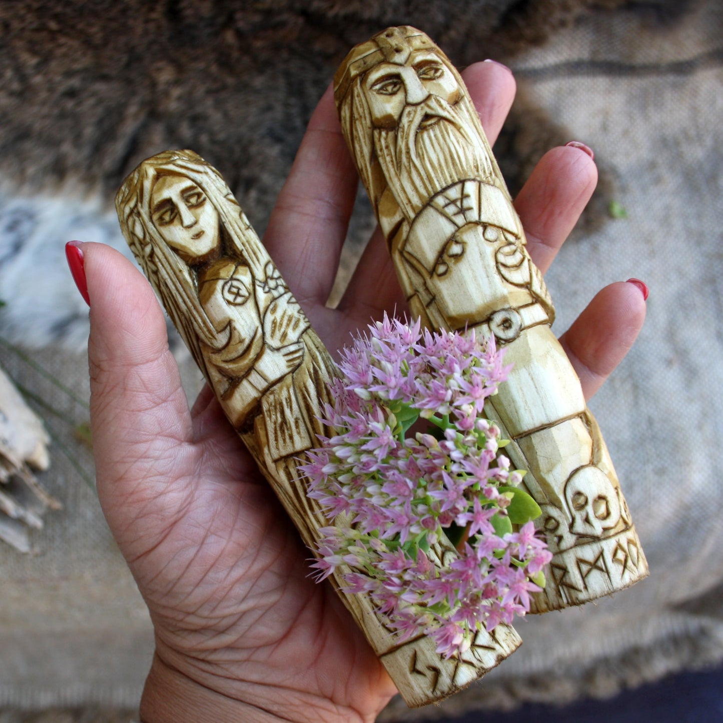 Hand holding Jord wooden statue with other Norse god hand carved figurines collection surrounded by pink flowers.