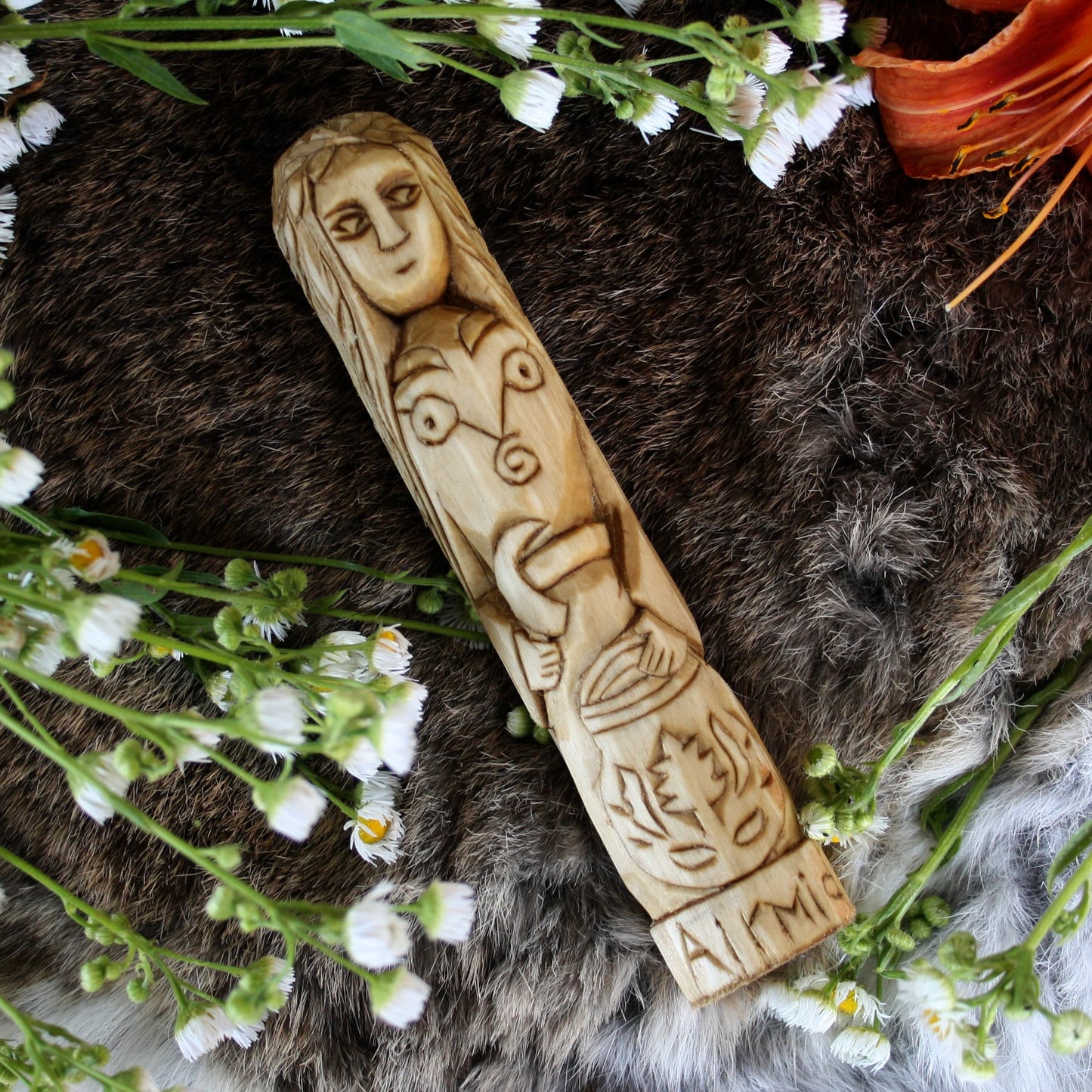 Airmid Celtic goddess displayed diagonally on a dark brown and gray fur surface surrounded by small white flowers.