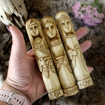 Norse Norns wooden statue set 15 cm Urd Verdandi Skuld held by a hand against a natural background with flowers.