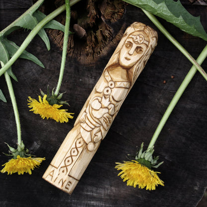 Áine Celtic goddess displayed on a dark wooden surface with green leaves and yellow dandelions.