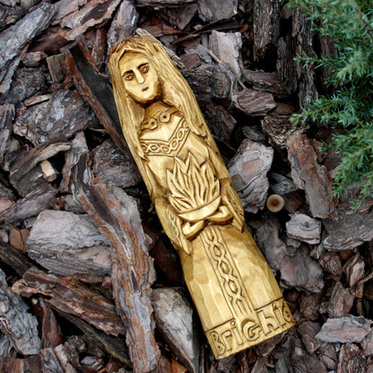 Hand-carved Brigid wooden statue displayed on bark in a natural outdoor setting