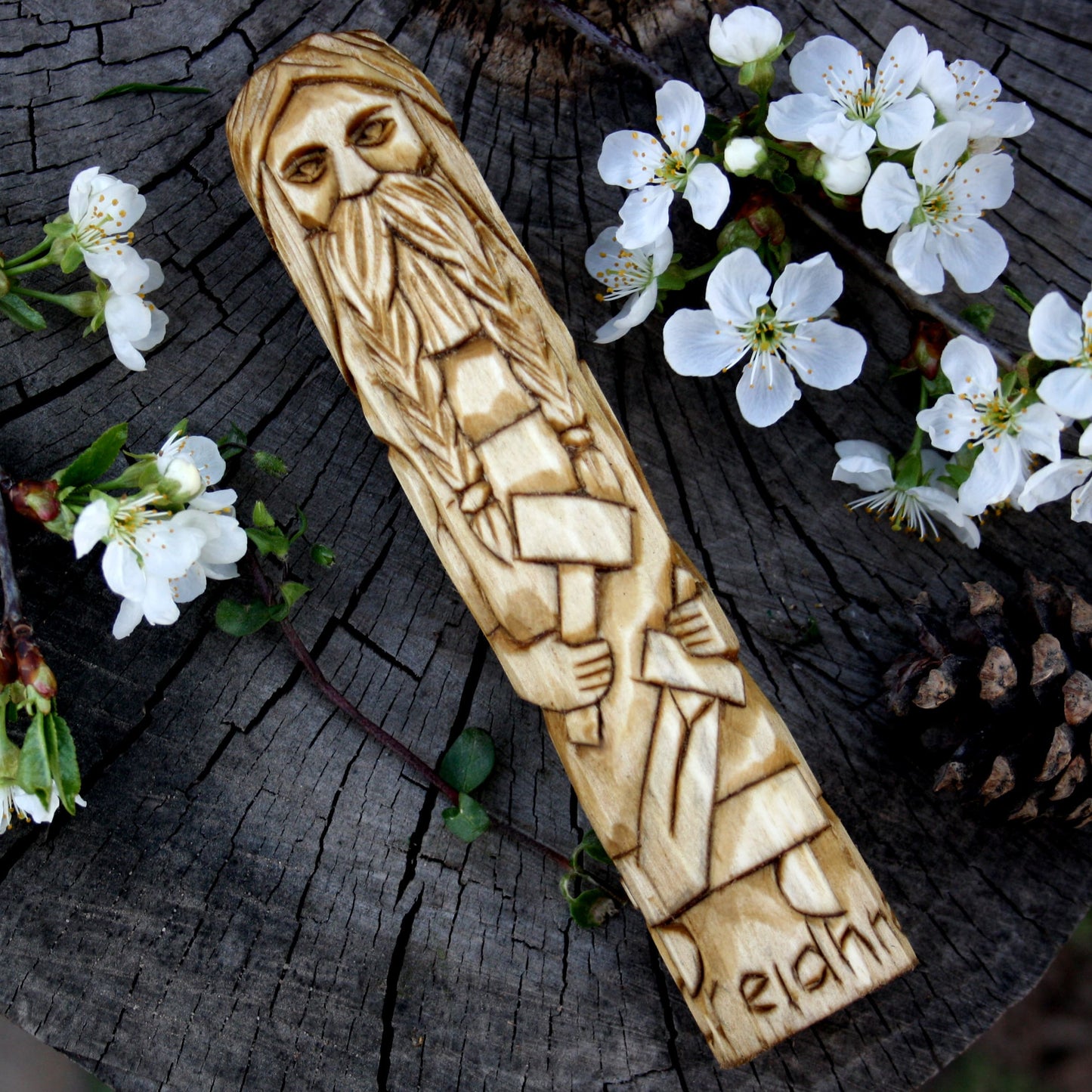 Creidhne Celtic god 15cm hand-carved wooden statue on a weathered tree stump with white blossoms and a pinecone around it.