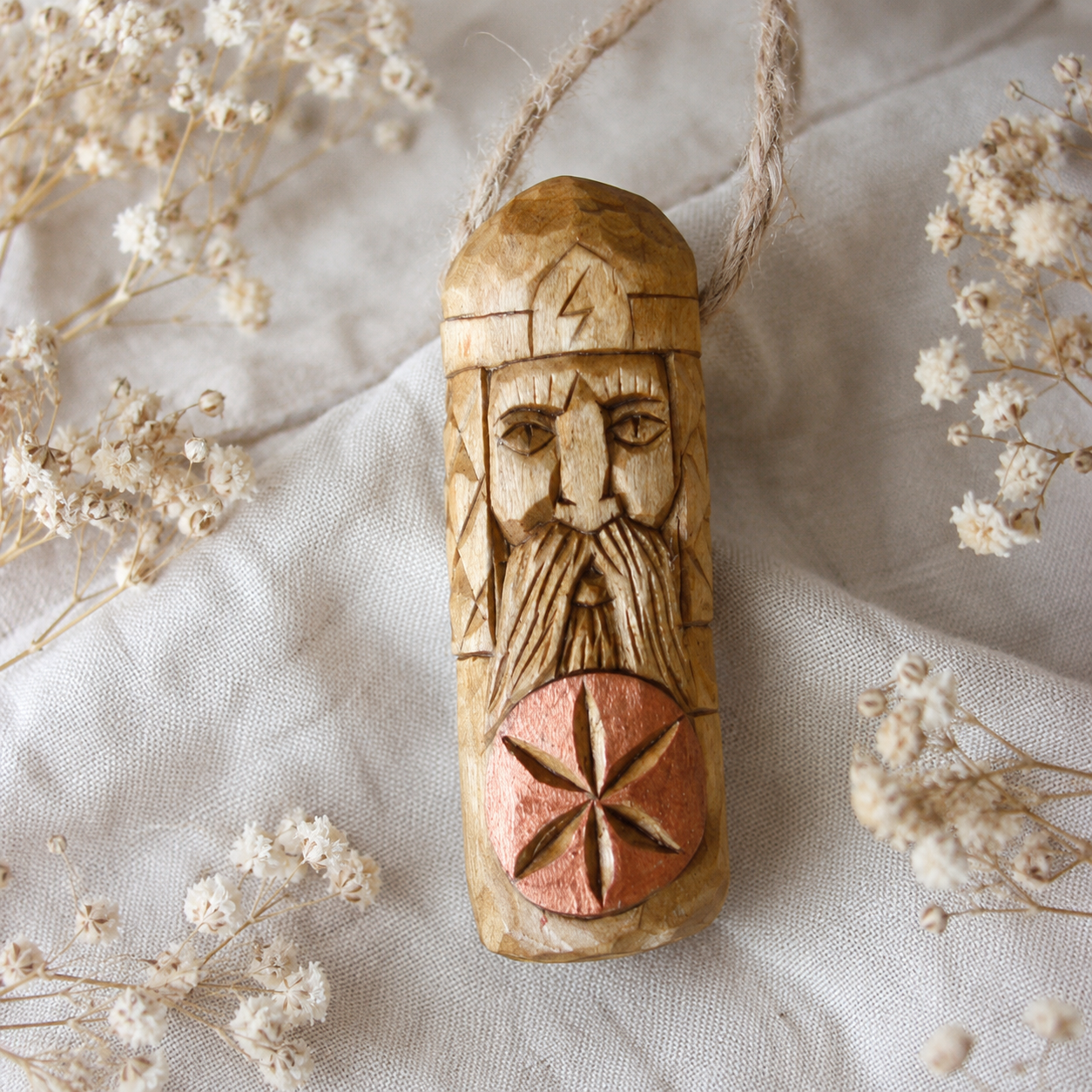 Hand holding a 10 cm Perun wooden amulet hanging from a twine string on a textured beige fabric with dried flowers around it.