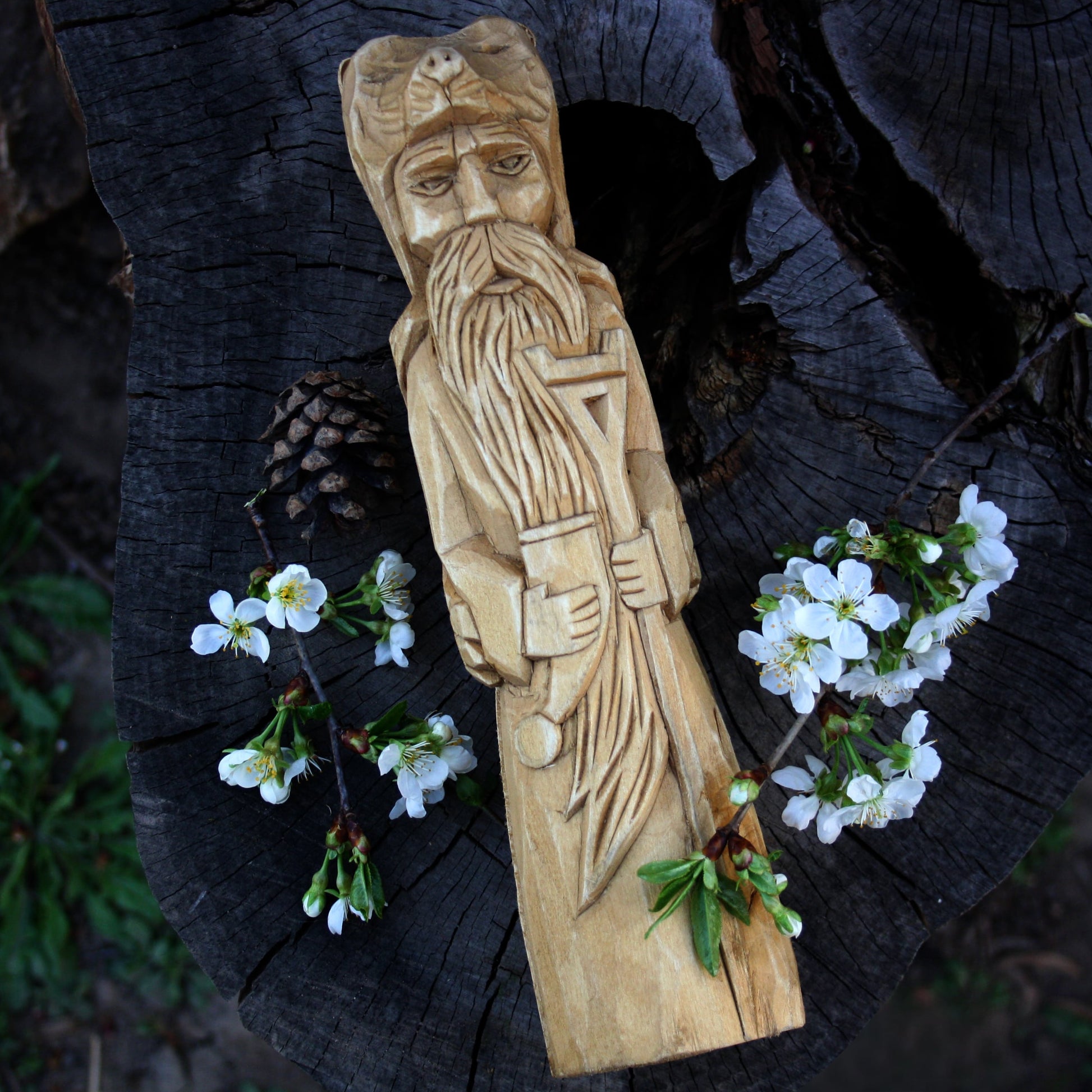 A hand-carved wooden statue of Slavic God Veles, displayed on a dark tree stump with small white flowers and a pinecone.
