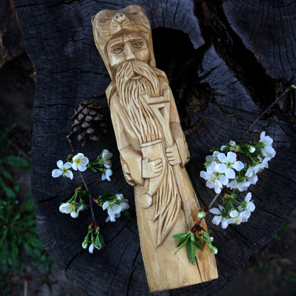 A hand-carved wooden statue of Slavic God Veles, displayed on a dark tree stump with small white flowers and a pinecone.